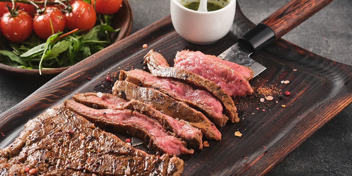 Sliced grilled beef steak on a wooden cutting board with tomatoes and sauce